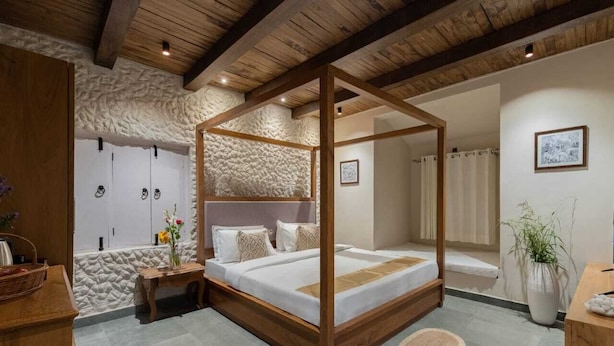 A four-poster bed with a stone wall backdrop, a wooden nightstand, and a potted plant. - AbhiRaj Villa (Udaipur)