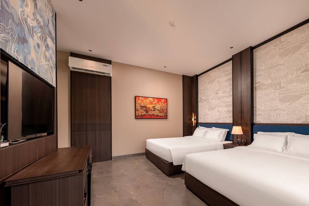A hotel room with two beds, a desk, a TV, an air conditioner, and a decorative wall.