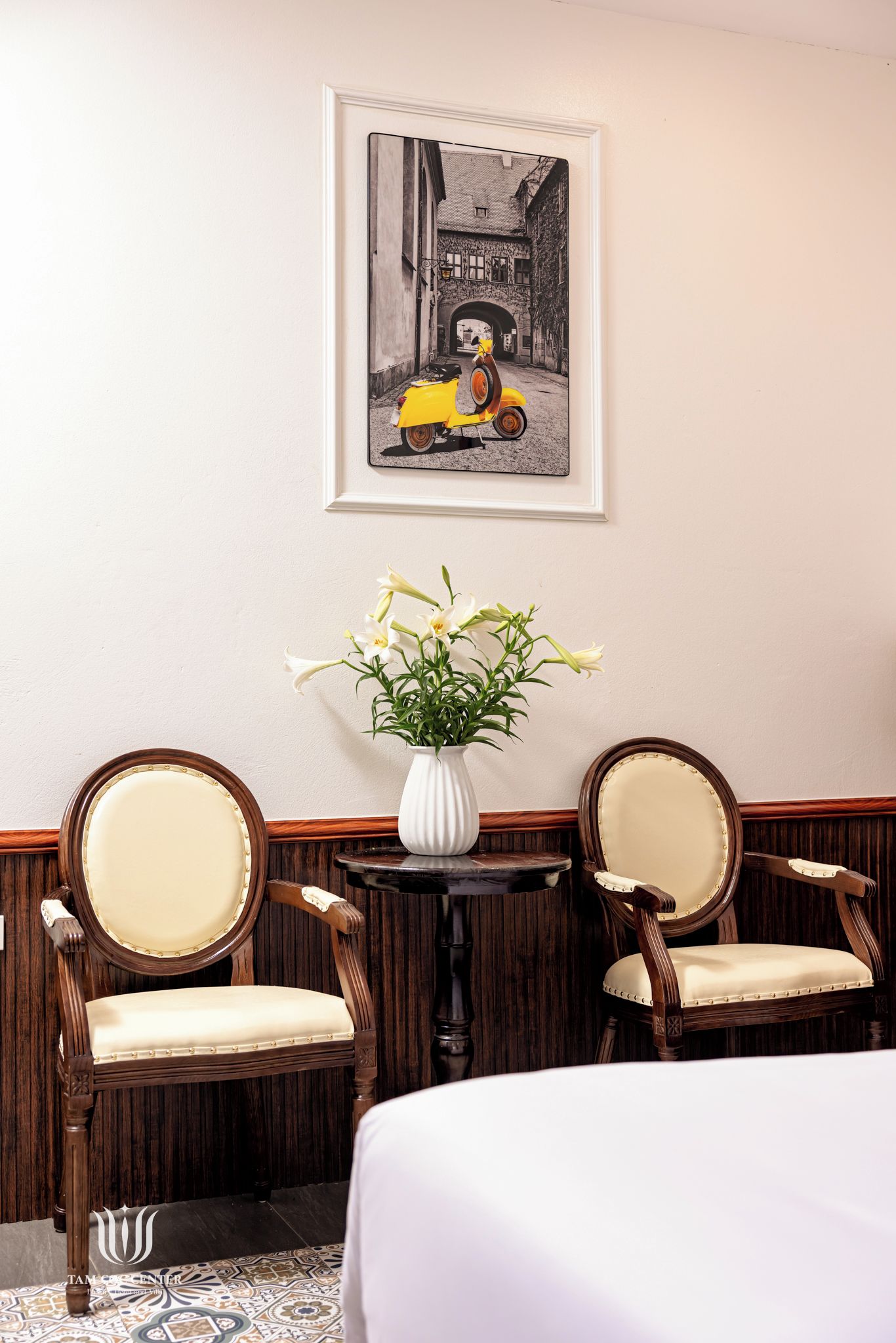 A hotel room with a bed, two upholstered chairs, a small table with a vase of flowers, and a framed picture on the wall.