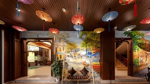 Entrada de un hotel con techo de madera, paraguas de colores, una silla colgante y un mural con una escena callejera.