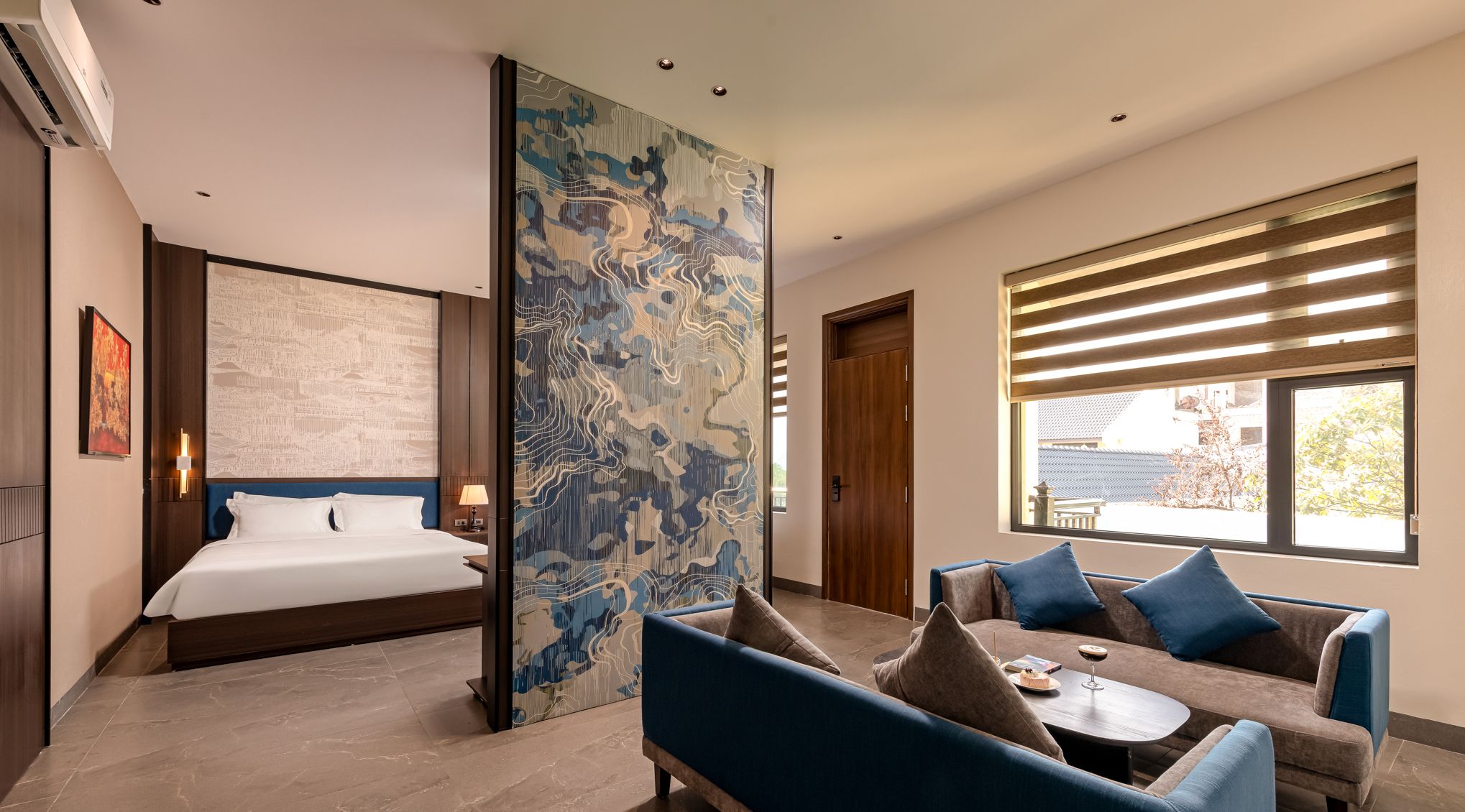 A hotel room with a bed, a sofa, a coffee table, and a large window with blinds.
