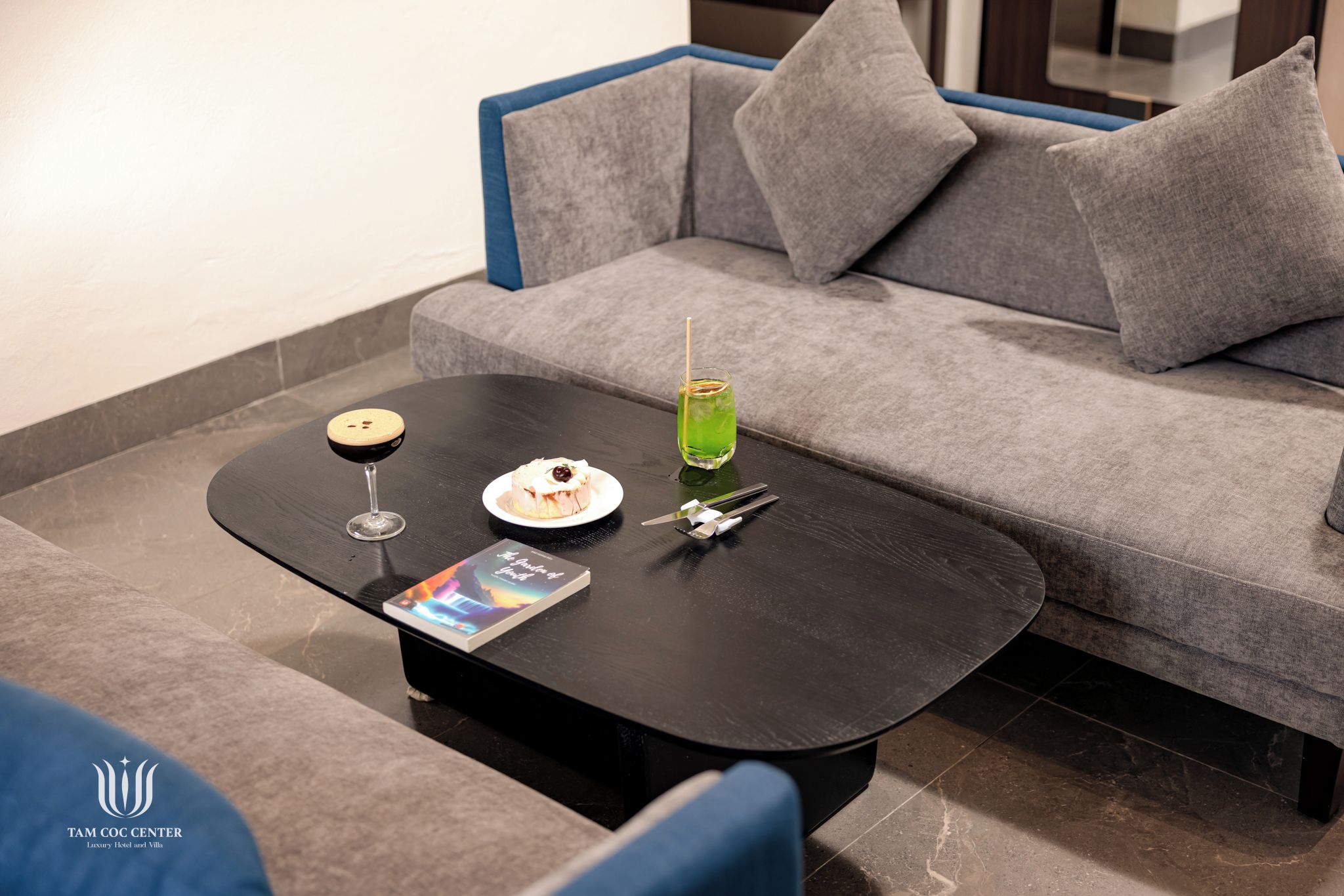 Een moderne loungehoek met een zwarte salontafel, een glas groene drank, een bord met een dessert en een wijnglas.