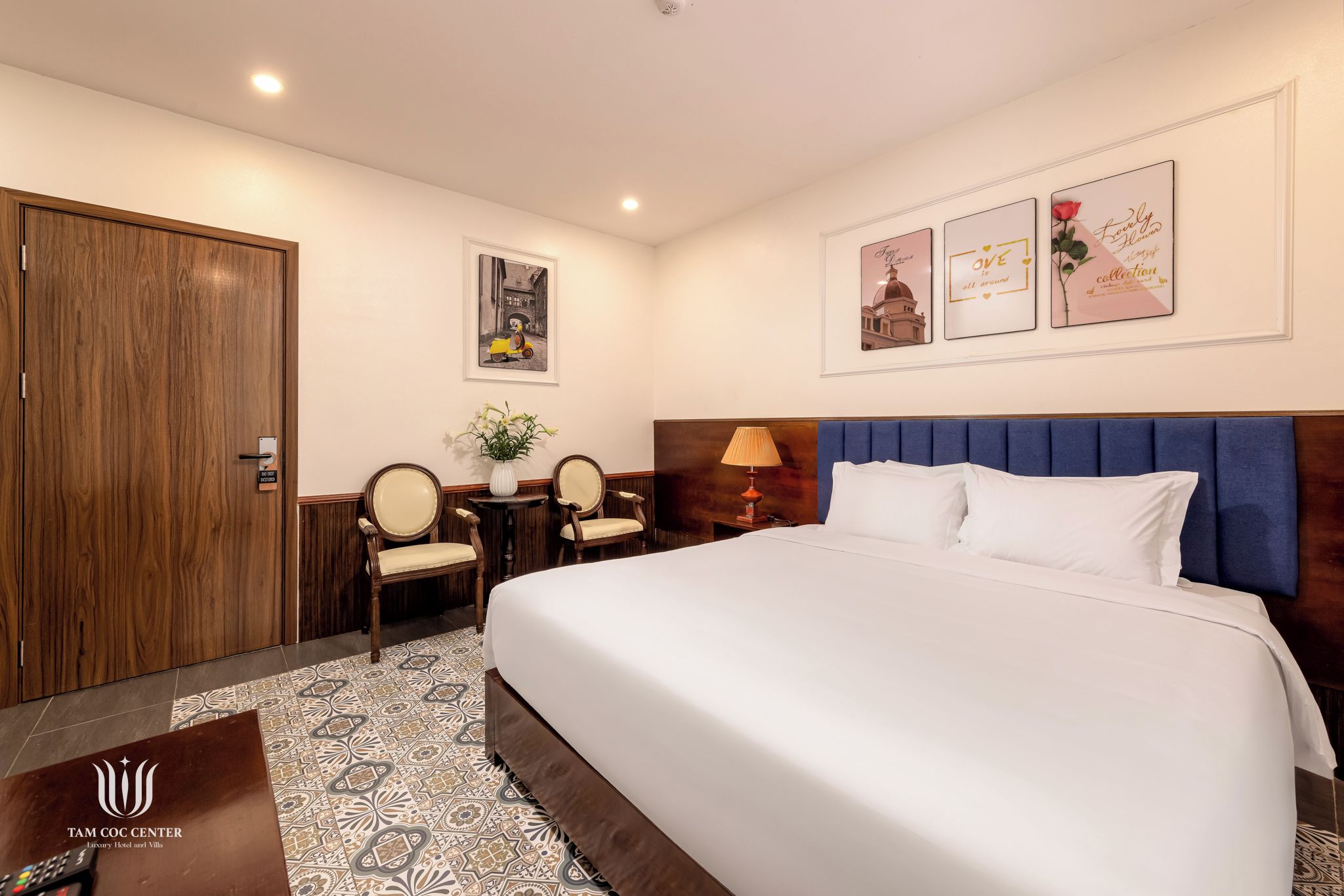 A hotel room with a large bed, a wooden wardrobe, a desk with a chair, and a small table with a lamp.