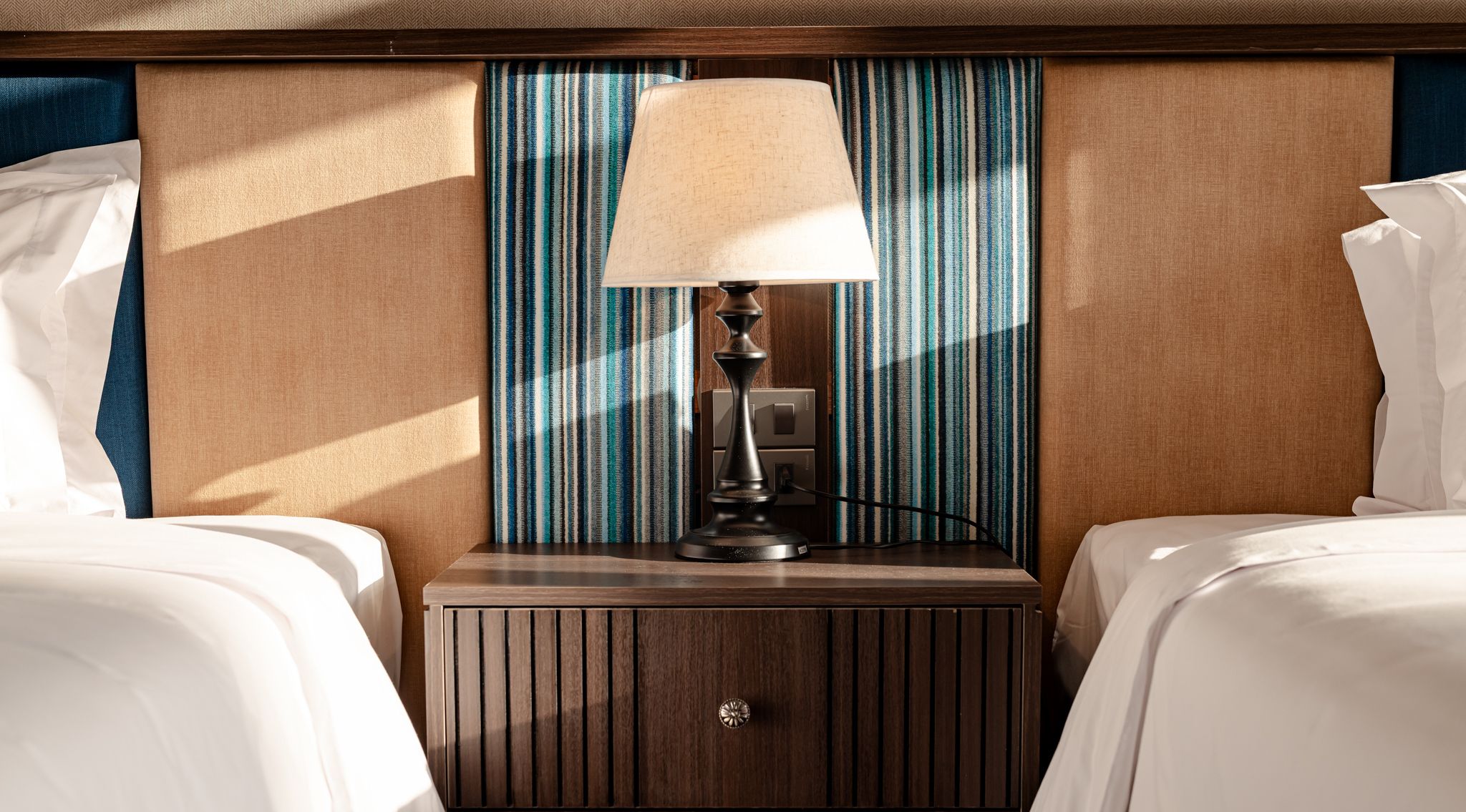 A hotel room with two beds, a nightstand with a lamp, and a striped curtain.