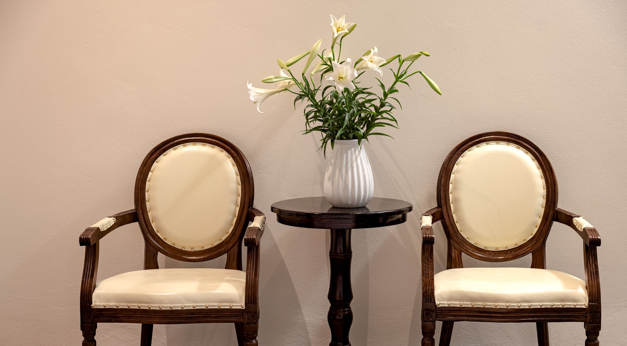 Two upholstered chairs with wooden frames and a round table with a white vase containing white flowers.