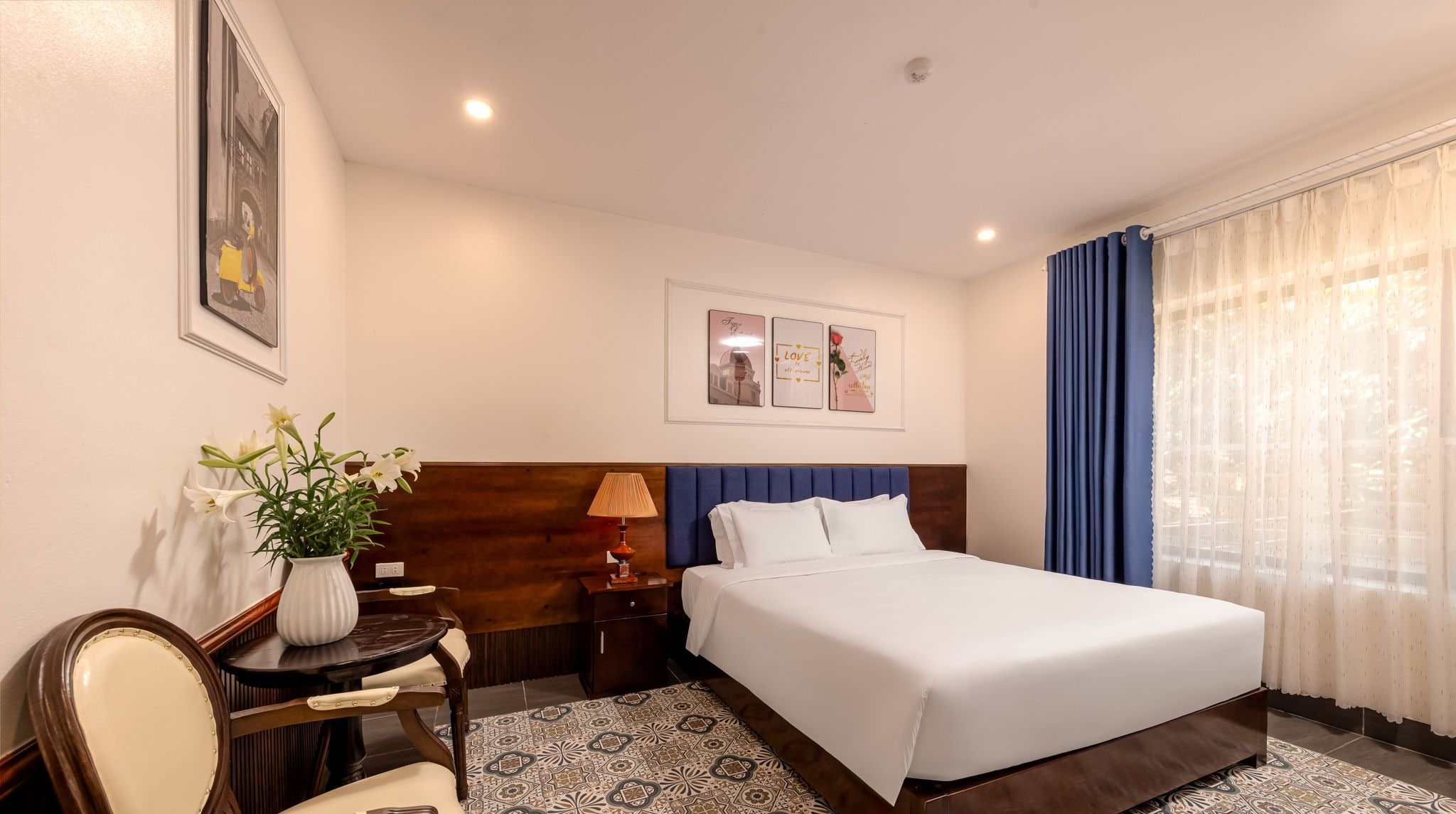 A hotel room with a large bed, a bedside table with a lamp, a chair, a vase with flowers, and framed pictures on the wall.