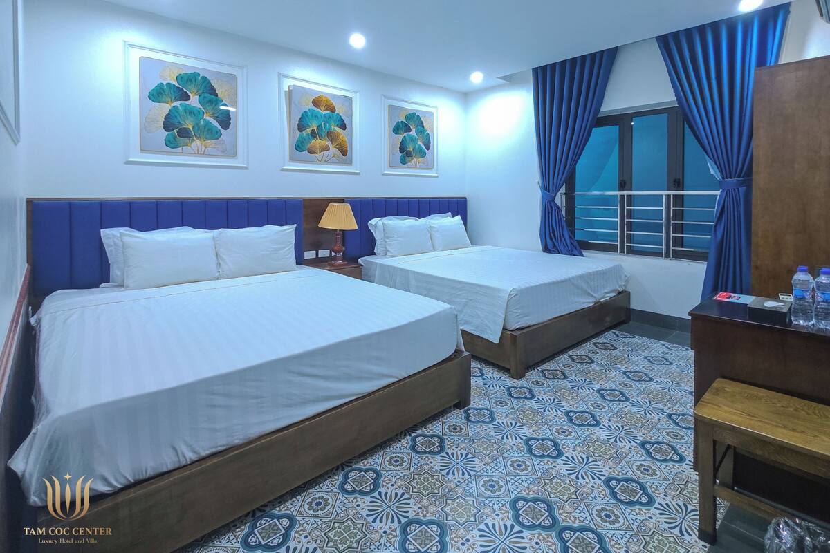 A hotel room with two beds, a nightstand, a lamp, and a view of the outside.