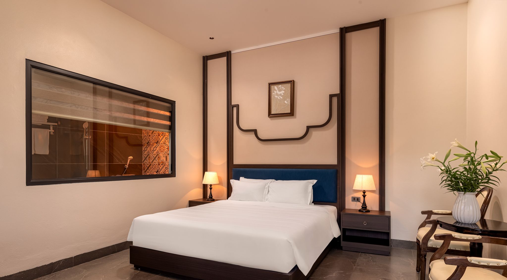 A hotel room with a large bed, two bedside tables, a nightstand, a wall-mounted picture, and a window with blinds.