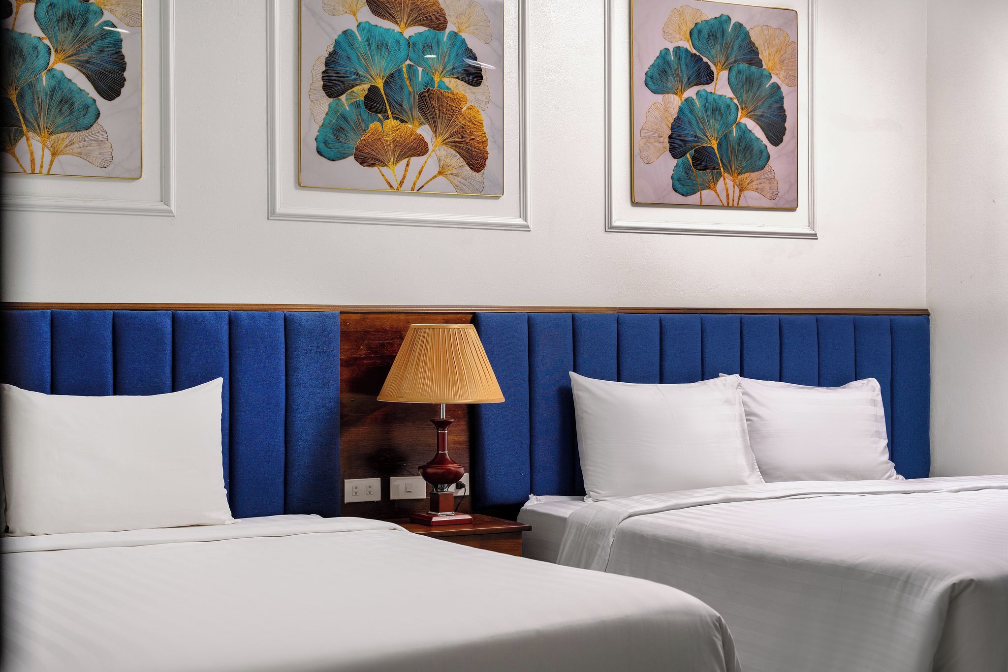 Camera d'albergo con due letti, una testiera blu, una lampada da comodino e opere d'arte incorniciate al muro.