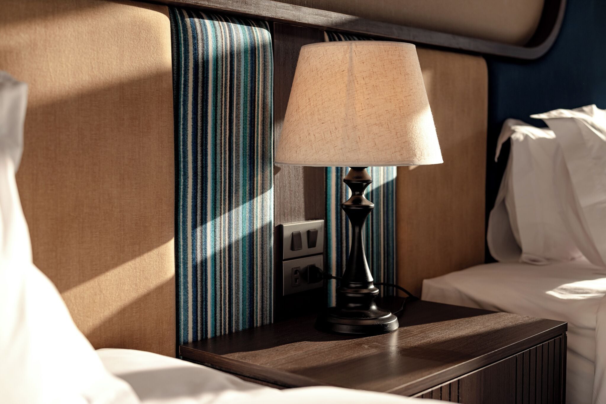 A neatly made bed with white linens, a bedside table with a lamp, and a striped curtain.