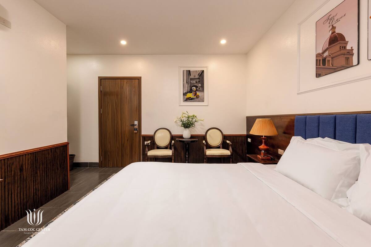 A hotel room with a large bed, two chairs, a small table with a plant, a lamp, and a framed picture on the wall.