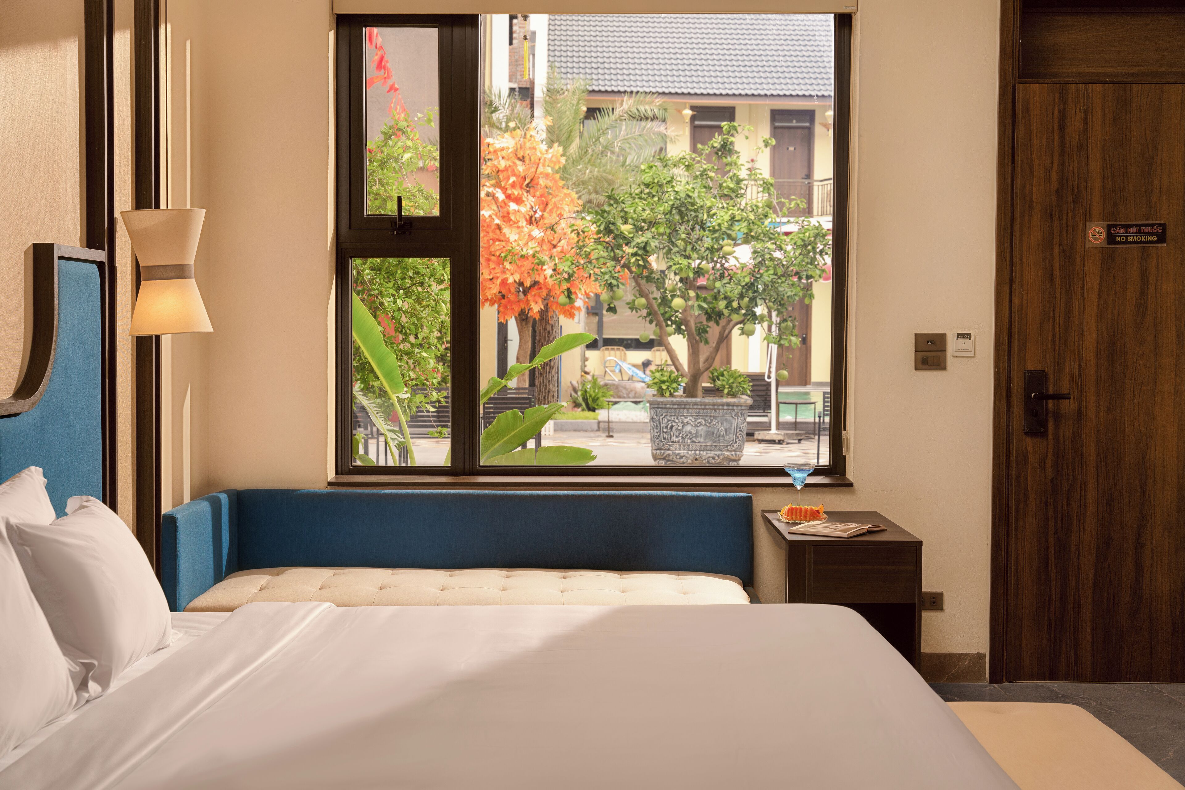 Habitación de hotel con cama, mesita de noche, lámpara, ventana que da a vistas del exterior y puerta.