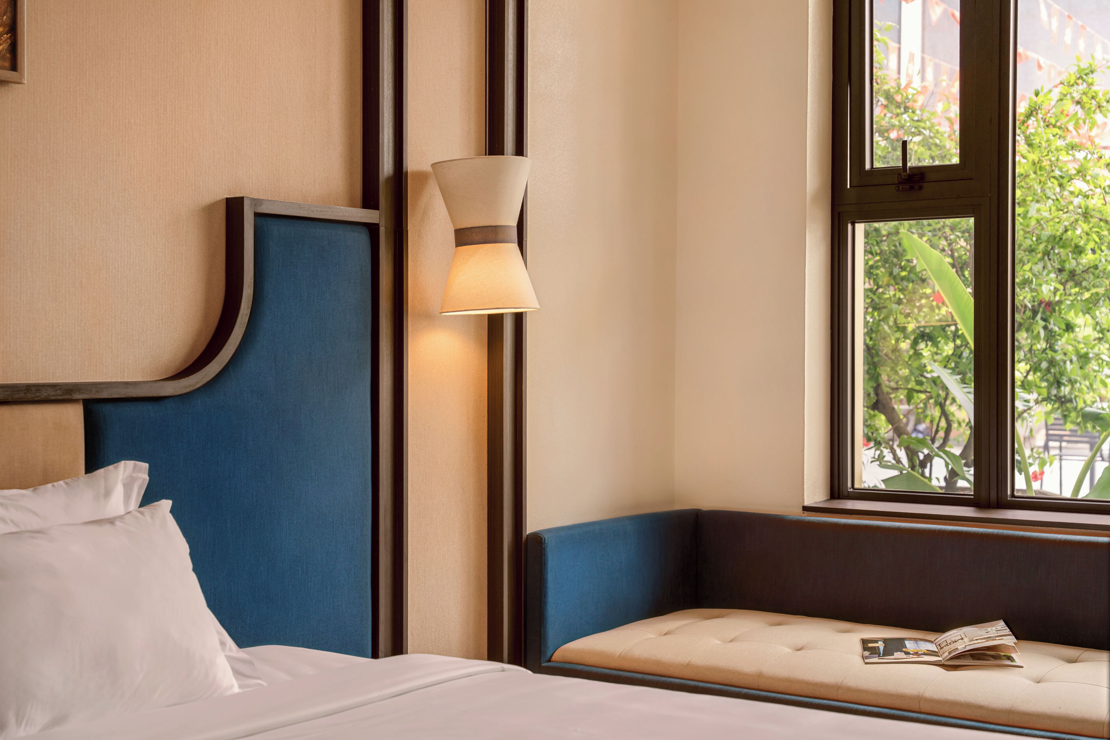 A hotel room with a bed, a blue headboard, a bedside lamp, a window with a view of greenery, and a bench with a magazine.