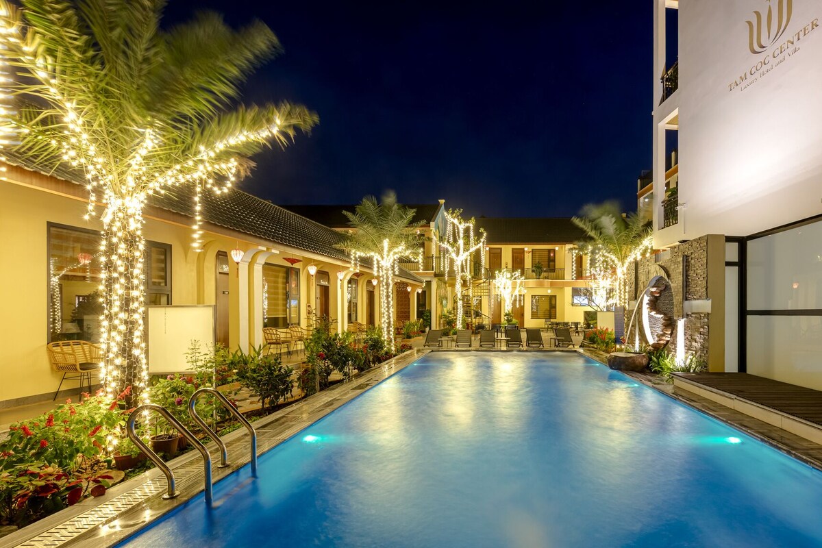 A swimming pool with lounge chairs and string lights, surrounded by buildings with a sign reading "TAM COC CENTER".
