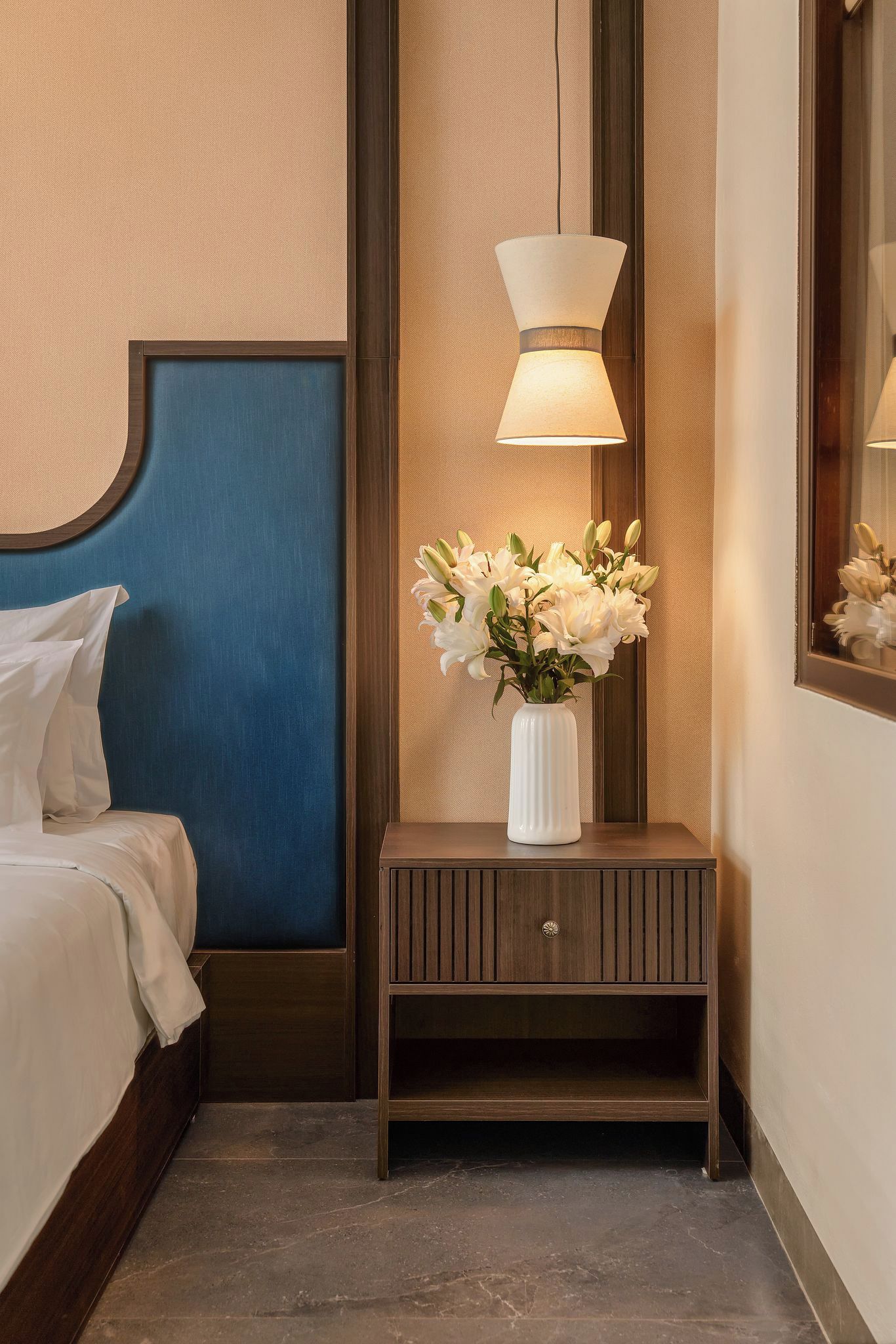 A hotel room with a bed, a nightstand with a vase of flowers, and a wall-mounted lamp.