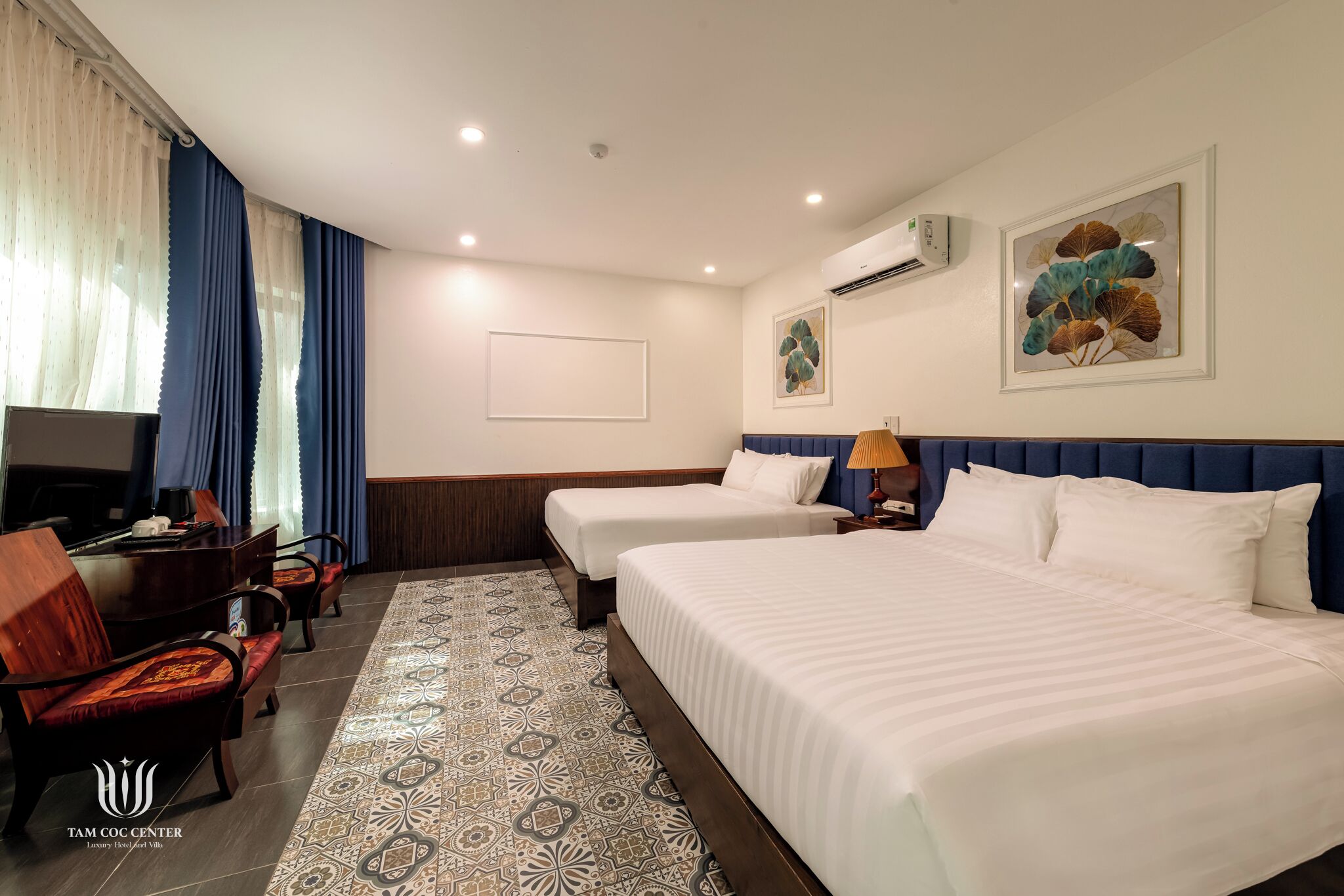 A hotel room with two beds, a desk with a chair, a TV, and a patterned rug.
