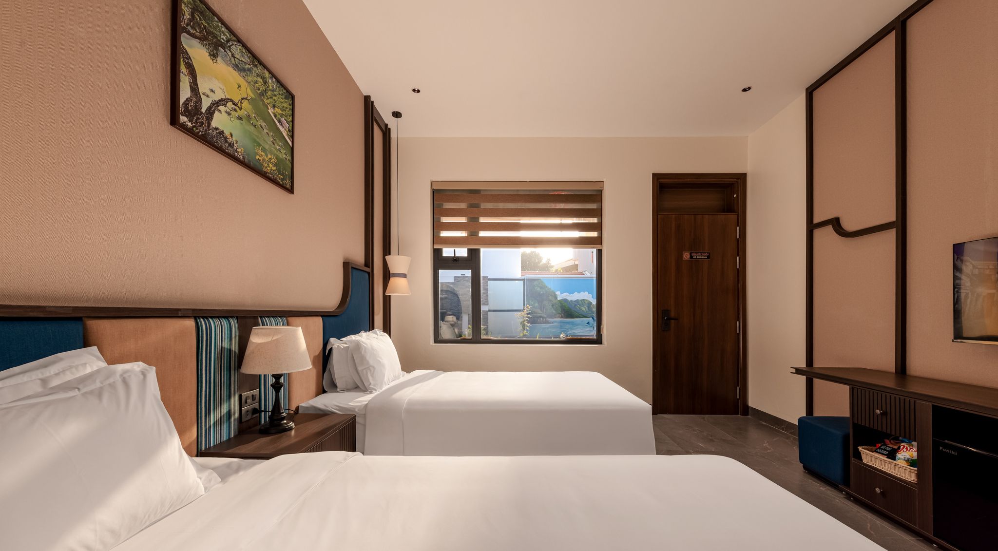 A hotel room with two beds, a window with a view, a painting on the wall, and a minibar.