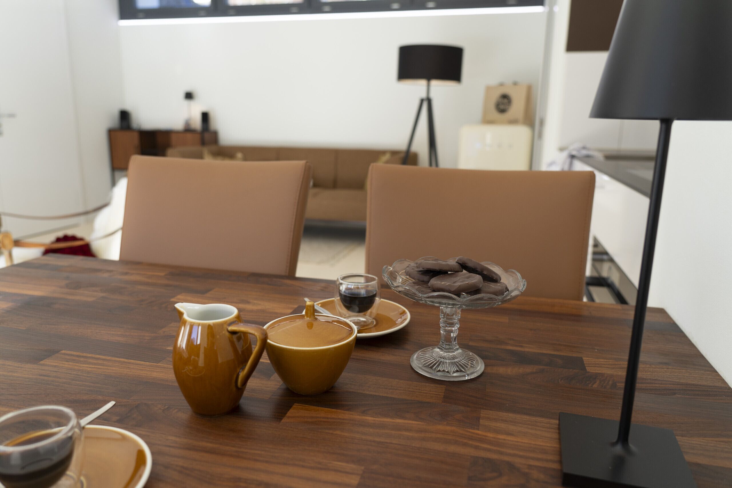 A wooden dining table with a coffee pot, cup, saucer, and a glass dish with cookies.