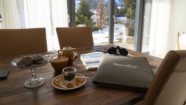 Una mesa de madera con un ordenador portátil, un plato de postre de vidrio, una cafetera, una taza y un cuaderno.