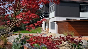 Una casa moderna de dos pisos con garaje, rodeada de un jardín ajardinado con follaje rojo y un césped bien cuidado.