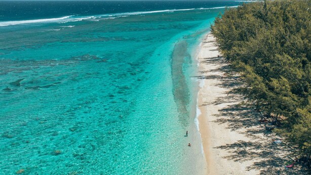Een strand met helder turquoise water, een zandstrand en weelderige groene begroeiing aan de rechterkant.