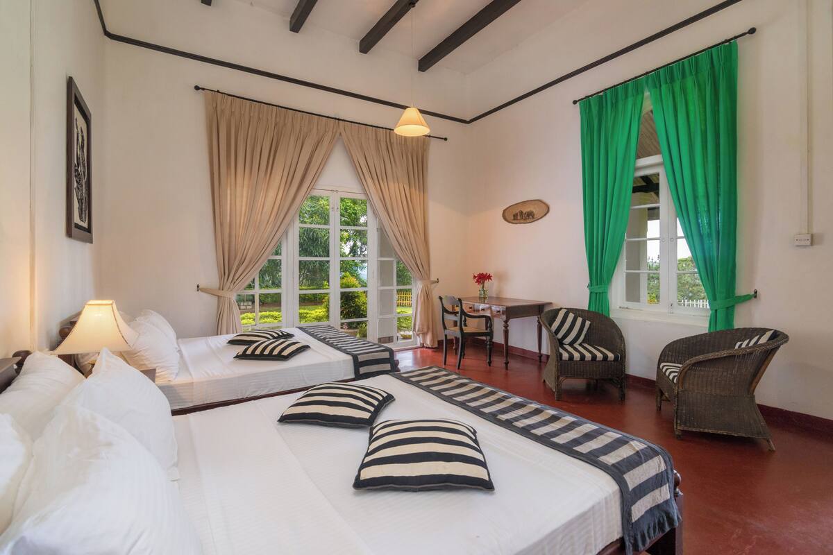 A hotel room with two beds, a desk, chairs, and a view of greenery outside.
