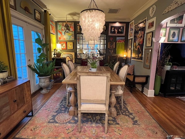 A dining area with a wooden table, chairs, and a chandelier. There are potted plants and framed pictures on the walls.