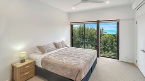 A modern bedroom with a large bed, a nightstand, and a view of greenery and a body of water through floor-to-ceiling windows. - The BoatHouse - Private 4 bedroom house (Whitsundays)