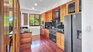 A modern kitchen with wooden cabinets, stainless steel appliances, and a large window offering a view of greenery. - The BoatHouse - Private 4 bedroom house (Whitsundays)