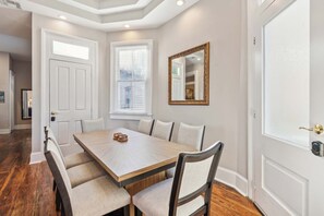 Une salle à manger avec une table en bois et six chaises, un grand miroir et une porte surmontée d’une fenêtre.