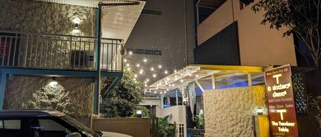 A multi-cuisine restaurant with outdoor seating, a car parked in front, and a sign indicating it's open for all.