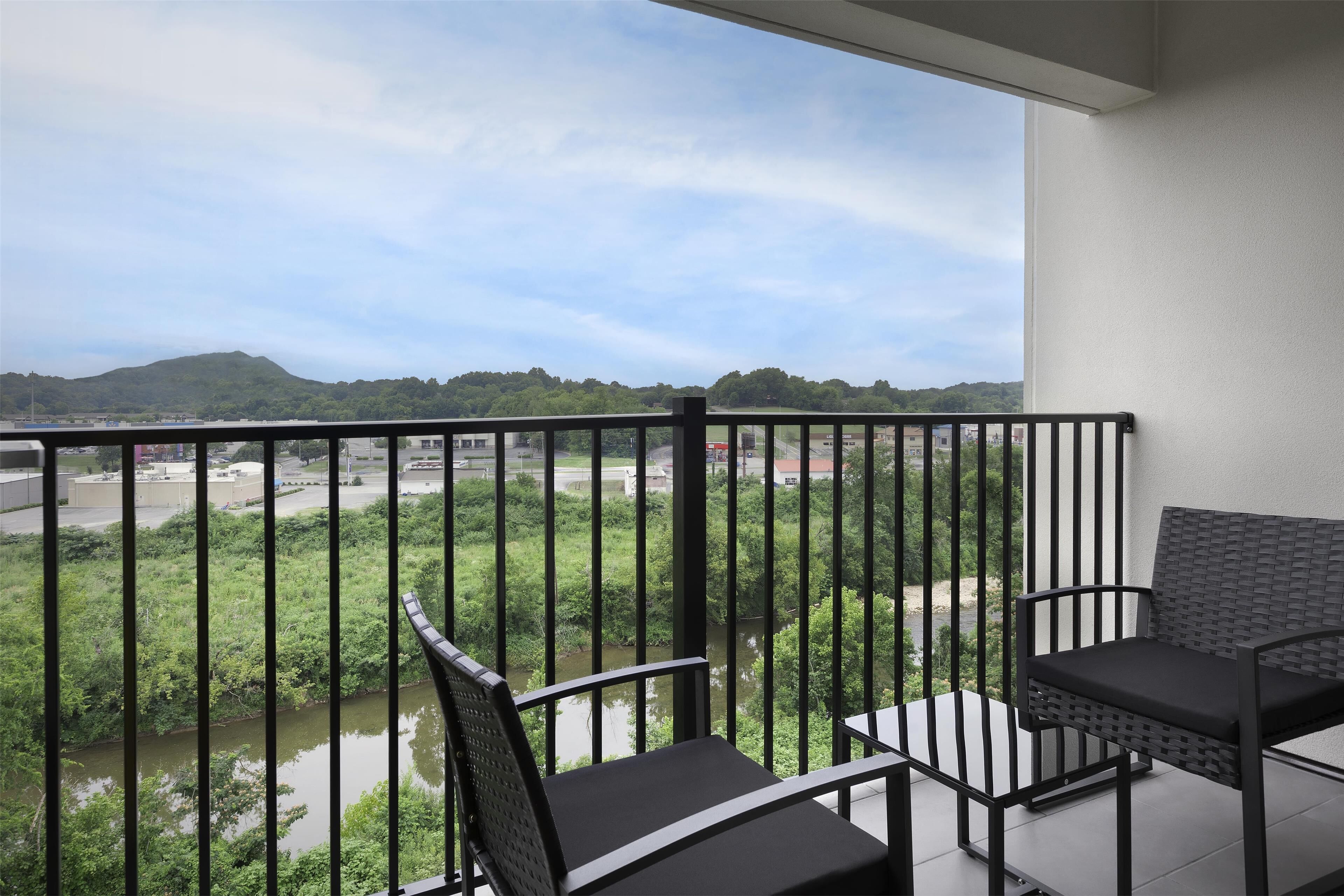 A balcony with a view of a river and hills, featuring two black chairs and a table.