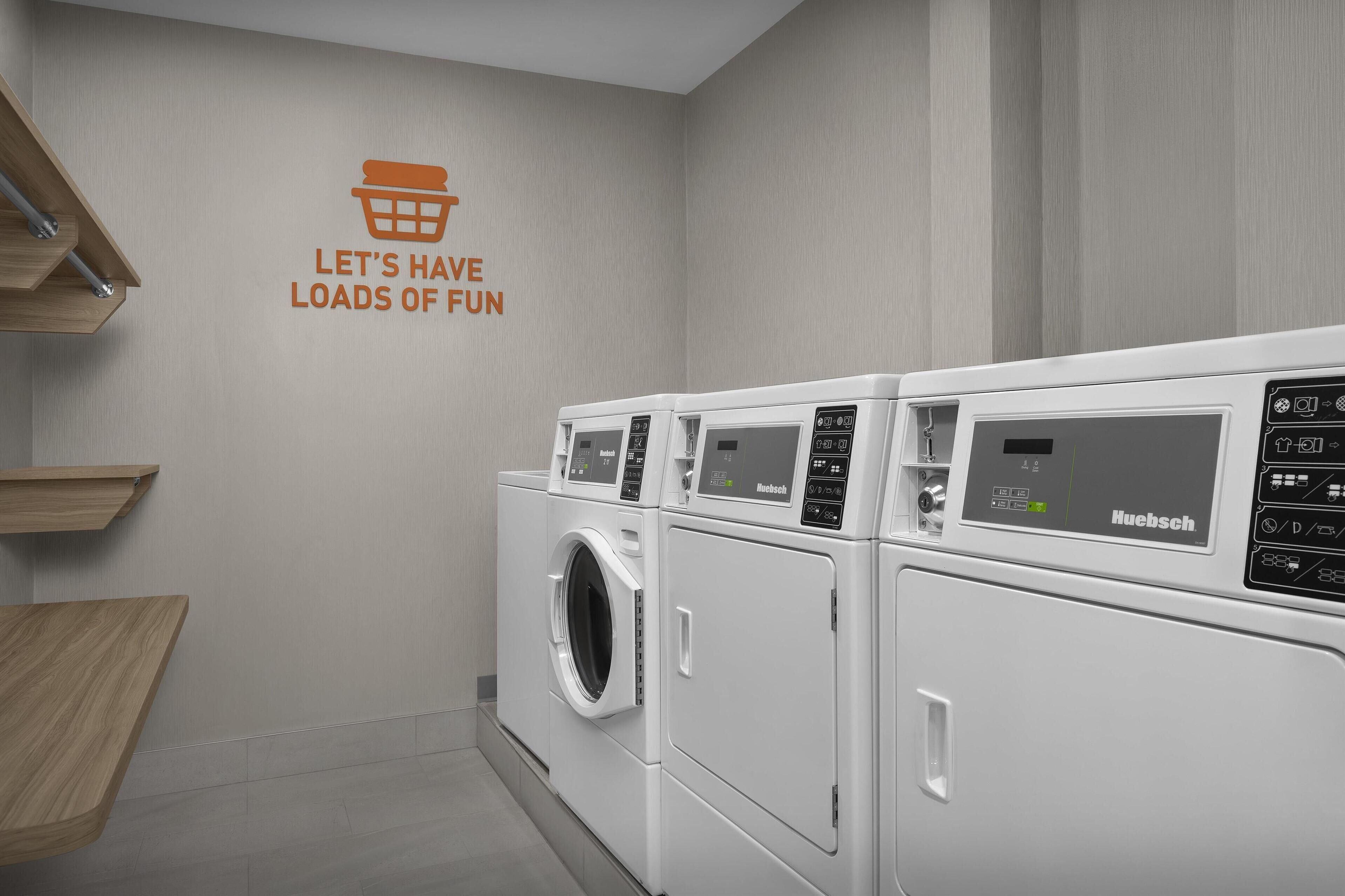 A laundry room with a wall-mounted sign that reads 'LET'S HAVE LOADS OF FUN' and a row of white washing machines.