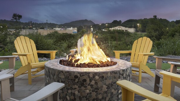 Un hogar de piedra con dos sillones Adirondack amarillos a su alrededor, situado en un patio con vistas a una colina y edificios al fondo.