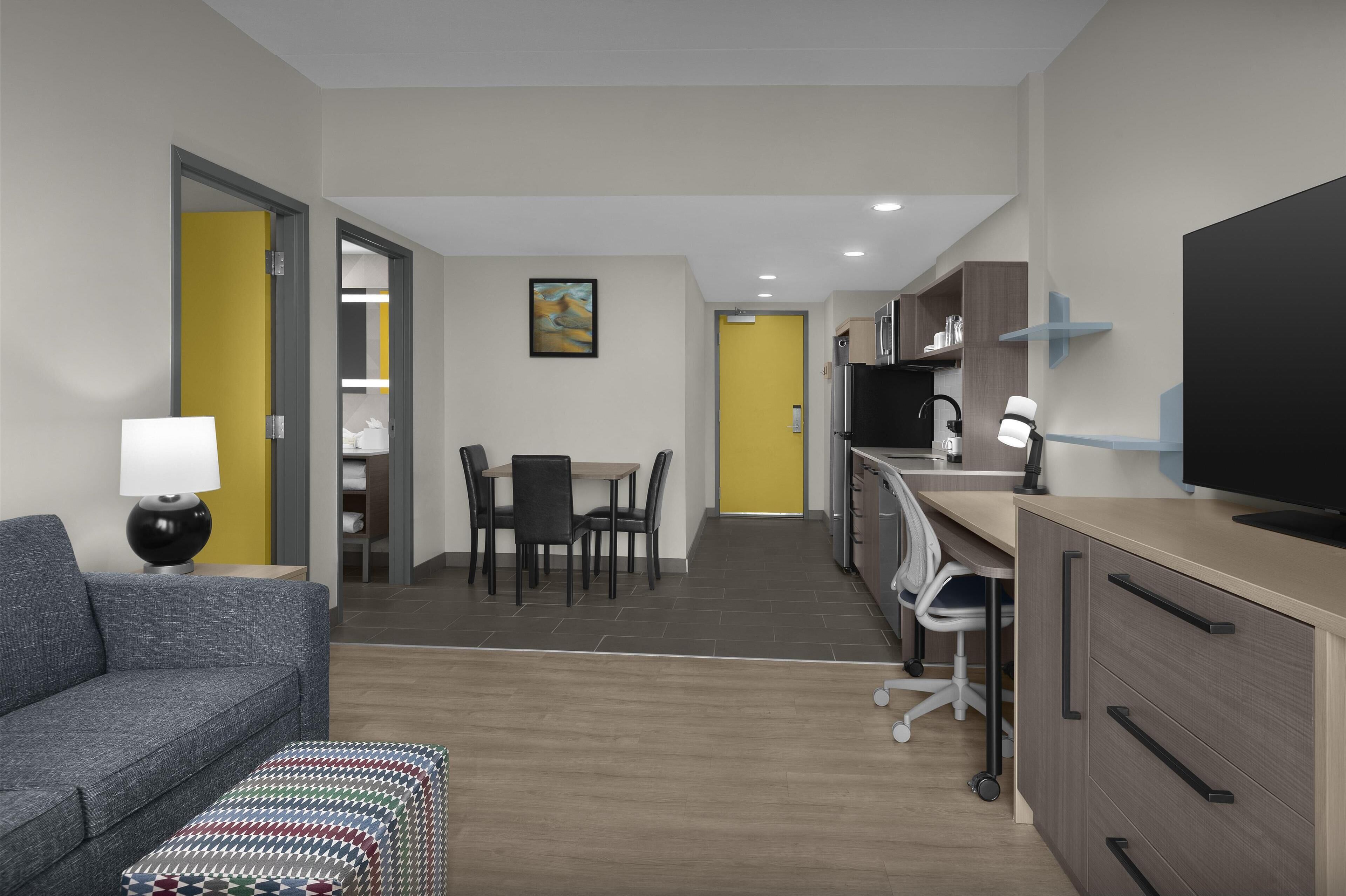 A modern hotel room with a grey sofa, a small table with a lamp, a dining area with chairs, a kitchen with a TV, and a yellow door.