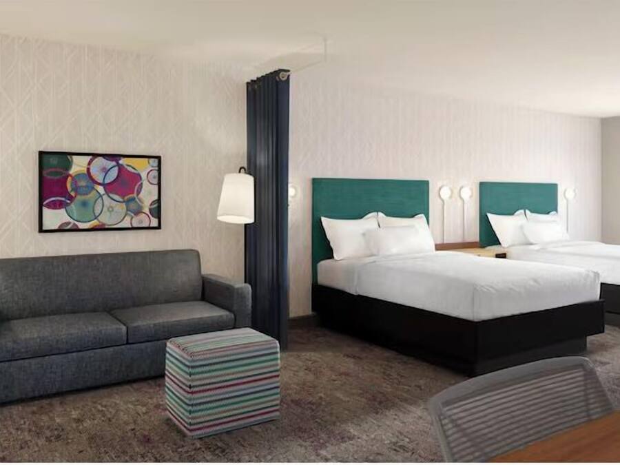 A hotel room with a gray sofa, two beds with teal headboards, a colorful abstract painting, a lamp, and a view of greenery through the window.