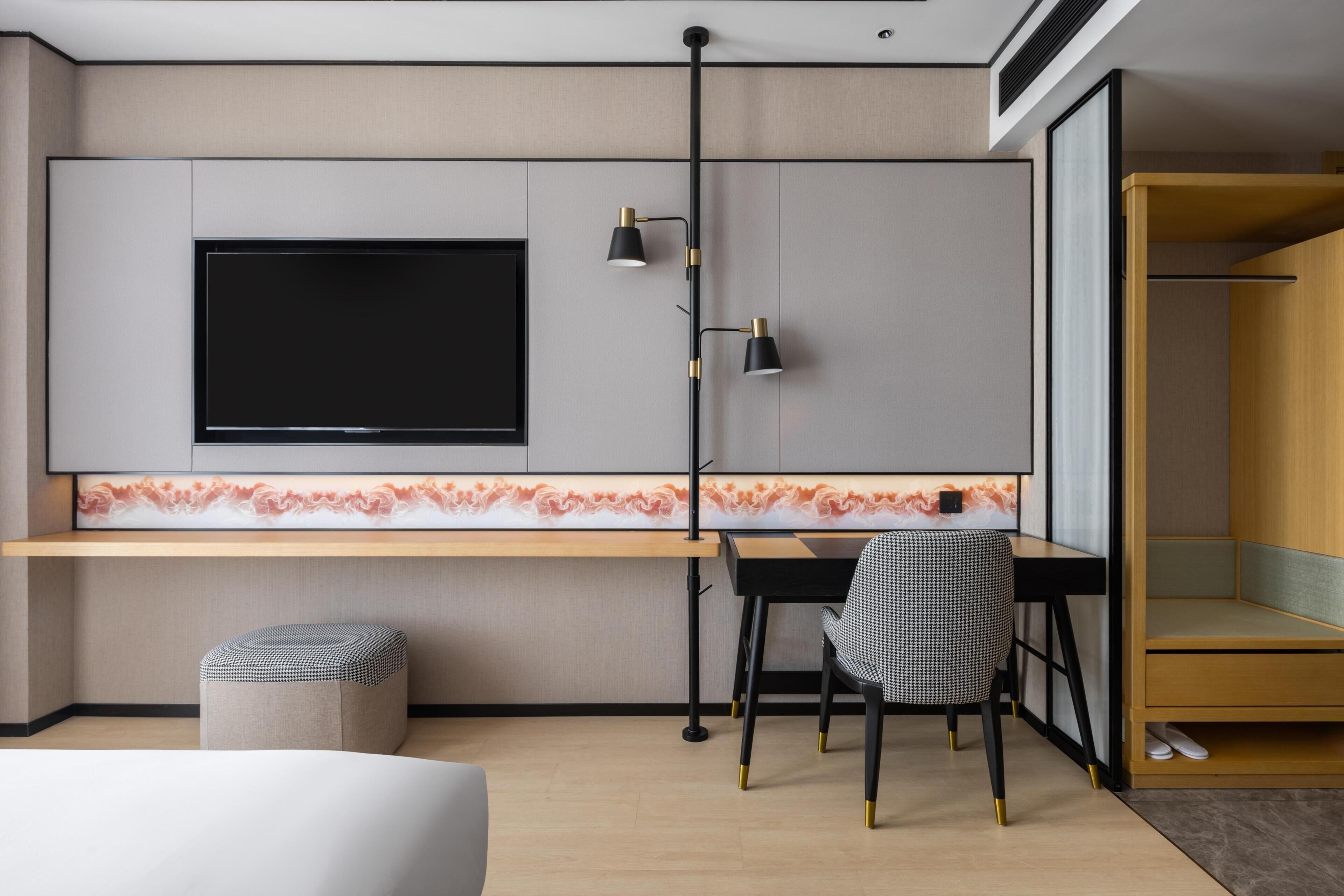 A modern hotel room with a flat-screen TV mounted on the wall, a desk with two lamps, a chair, and a small ottoman.