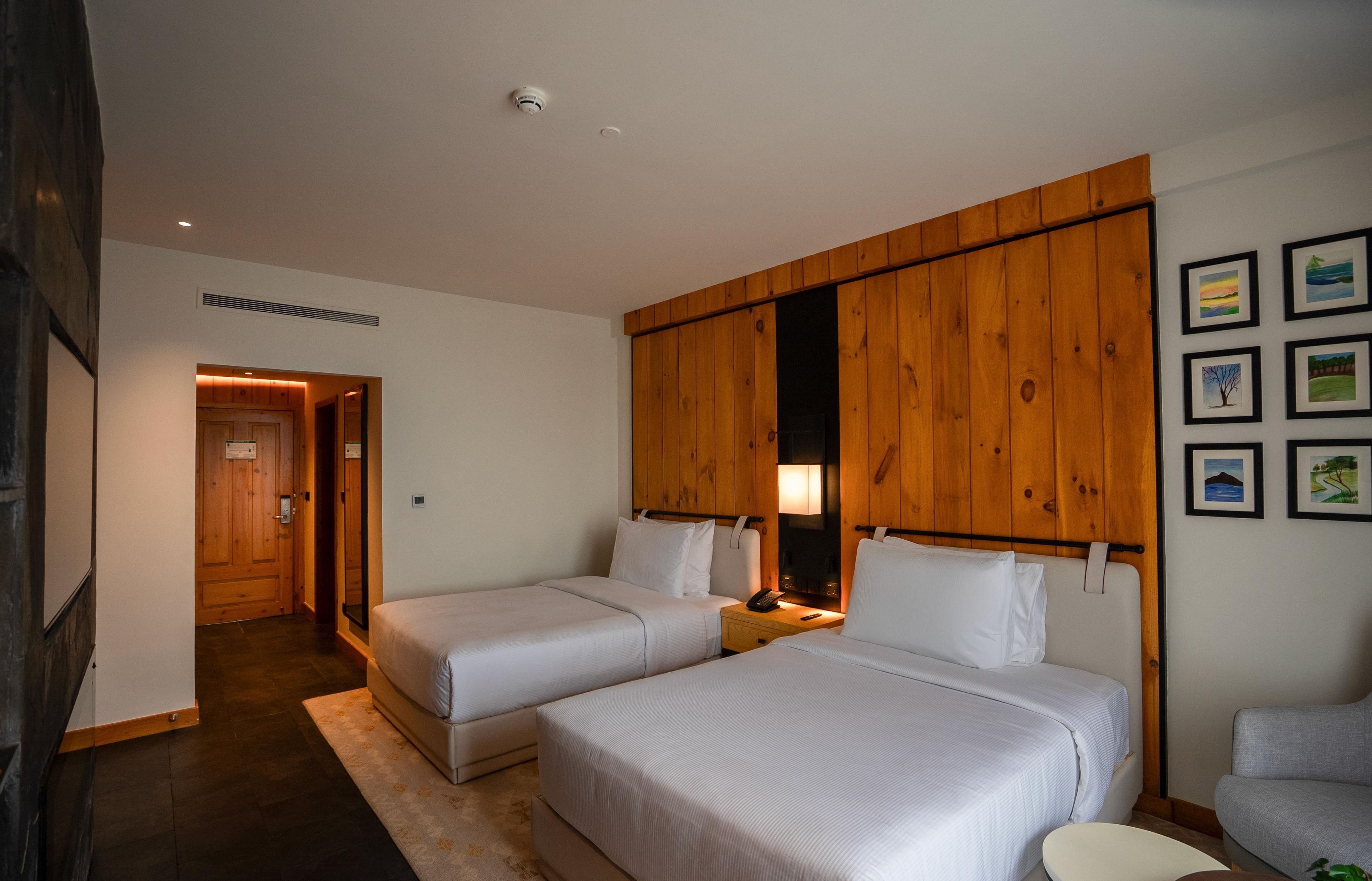 A hotel room with two beds, a wooden wardrobe, a nightstand with a lamp, and a small table.