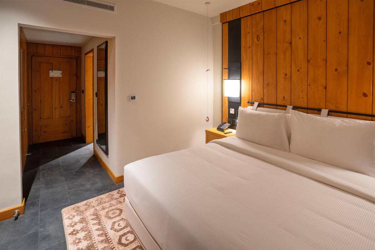 Een slaapkamer met een groot bed, een wand van houten panelen, een nachtkastje met een lamp en een vloerkleed met dessin.