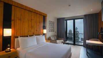 A hotel room with a large bed, a desk, a chair, a balcony with a table, and a view of the outdoors.