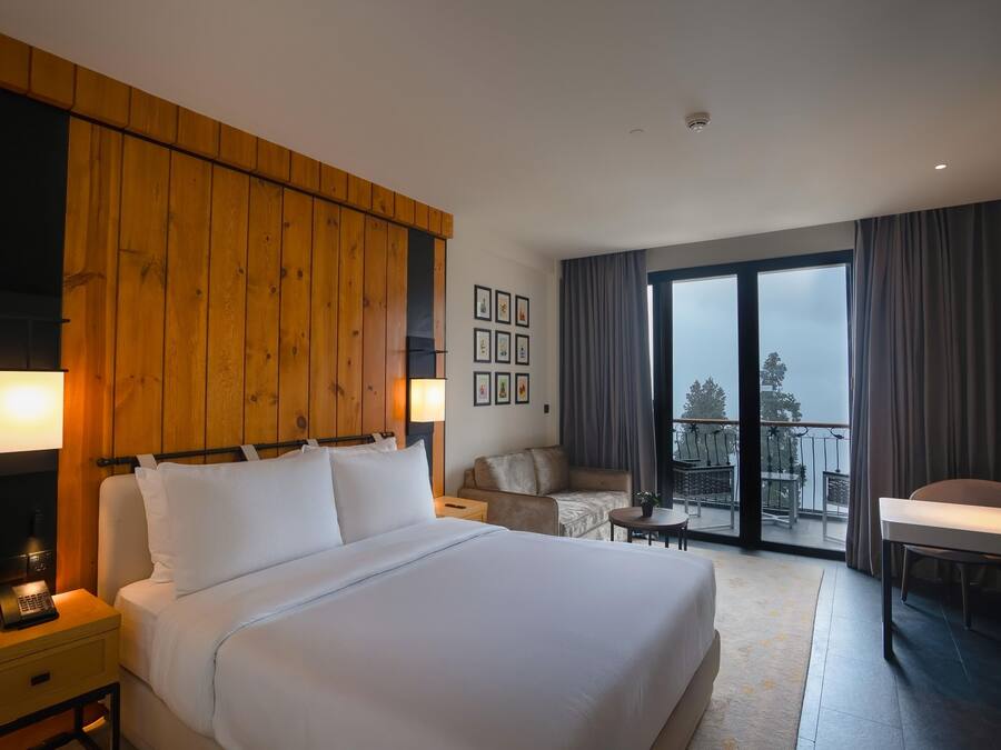 A hotel room with a large bed, a desk, a chair, a balcony with a table, and a view of the outdoors.