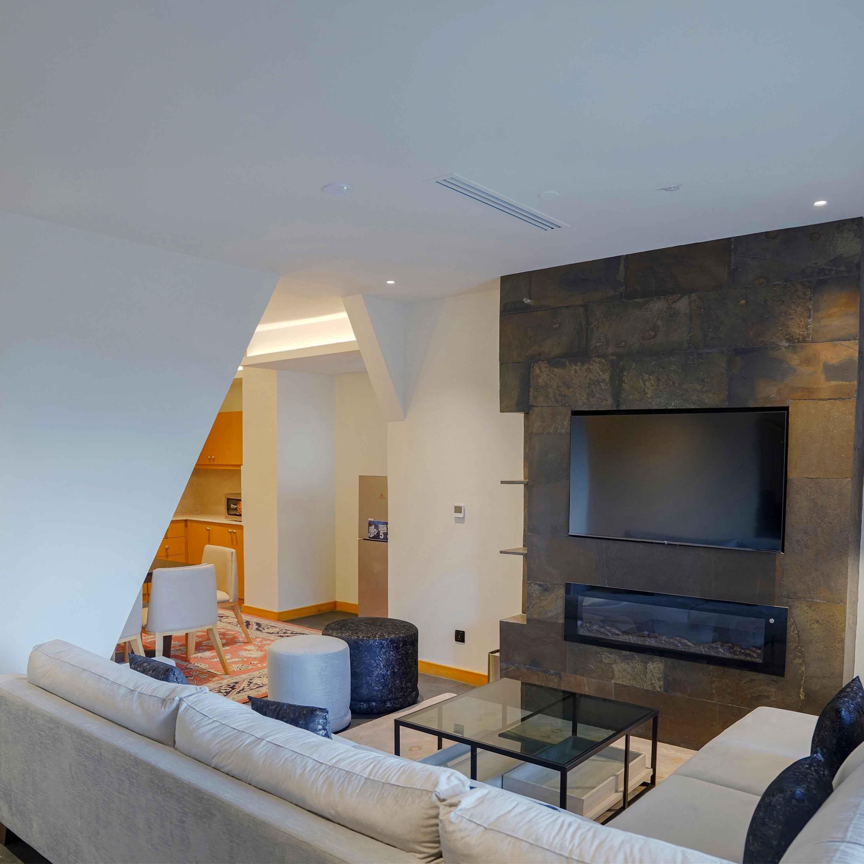 Una sala de estar moderna con un sofá, un reposapiés y una mesa de centro de vidrio. Un televisor de pantalla plana está montado en una pared de piedra.