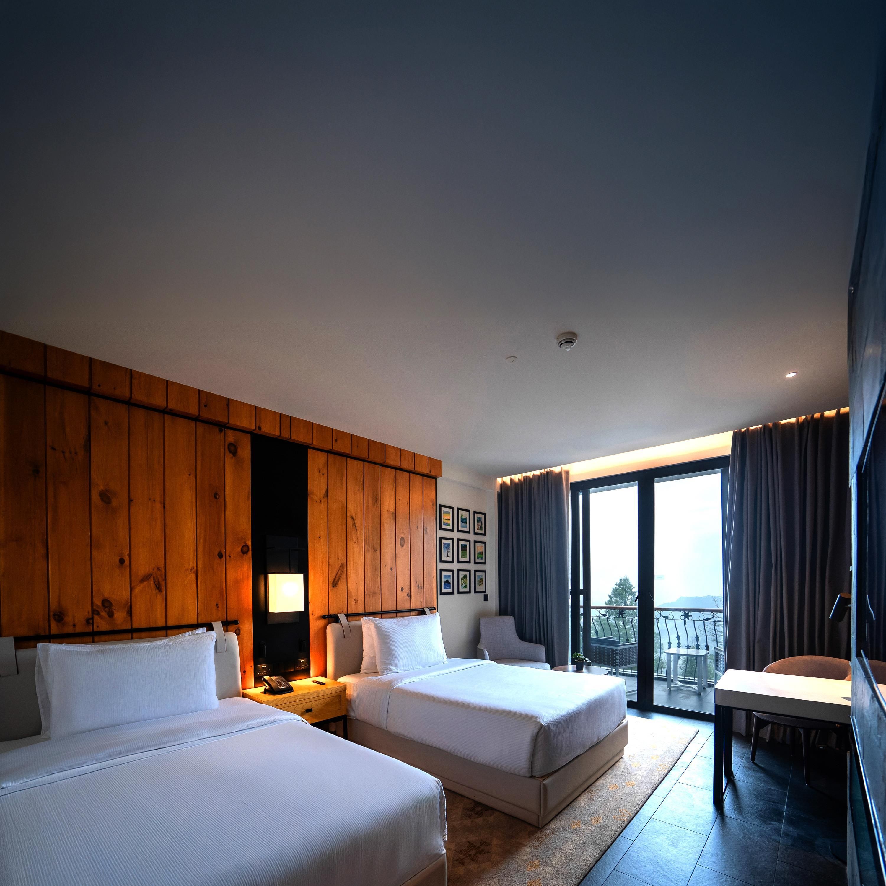 Quarto de hotel com duas camas, uma escrivaninha, uma cadeira, uma janela grande com cortinas e um painel de parede em madeira.