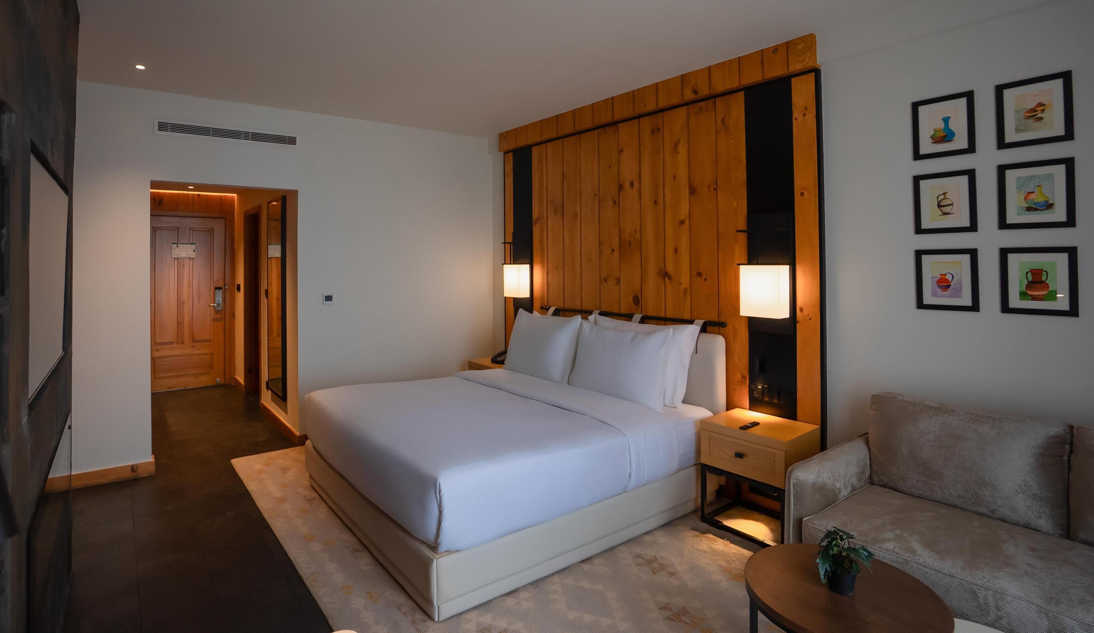 Habitación de hotel con una cama grande, cabecera de madera, mesita de noche con lámpara, un pequeño sofá y una pared con cuadros.