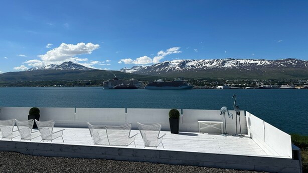 A deck with white chairs and a bench overlooking a body of water with cruise ships and mountains in the background. - Halllandsnes Apartments (Akureyri)