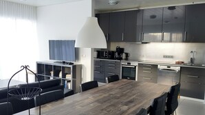 A modern kitchen with dark cabinets, a wooden dining table, and a large window with curtains. - Halllandsnes Apartments (Akureyri)