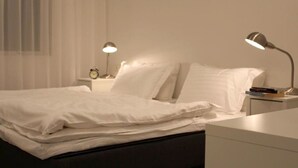 A neatly made bed with white linens, two bedside lamps, a small bedside table with a clock, and a white nightstand. - Halllandsnes Apartments (Akureyri)