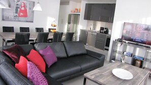 A modern living room with a black leather sofa, a dining area with a wooden table and chairs, a kitchen with dark cabinets, and a flat-screen TV on a stand.