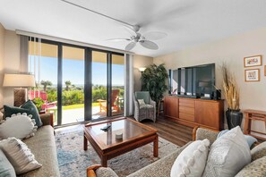 Un salon spacieux doté d’une grande porte-fenêtre coulissante donnant sur un balcon, d’un ventilateur de plafond, d’une télévision et d’un coin repas avec des meubles d’extérieur.