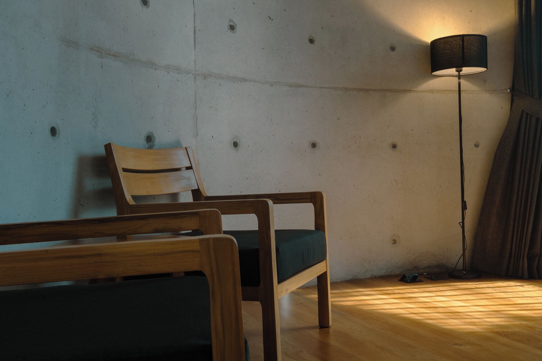 Ein Zimmer mit einem Holzstuhl, einer Stehlampe und einer Betontwand mit sichtbaren Holzstielen.